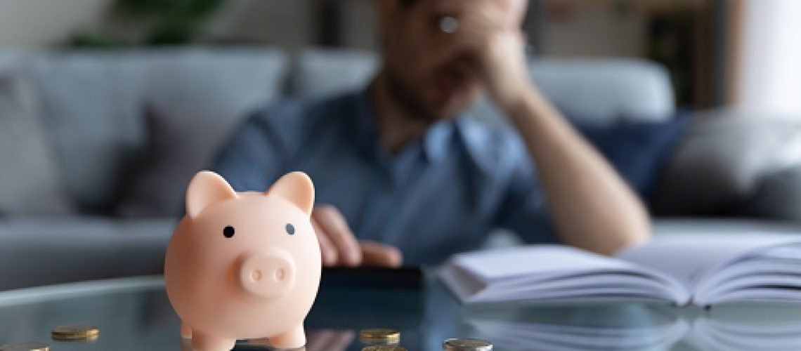 Close up focus on piggybank on table, young man feeling stressed calculating expenditures or taxes, managing future payments or planning investments, suffering from lack of money, accounting concept.