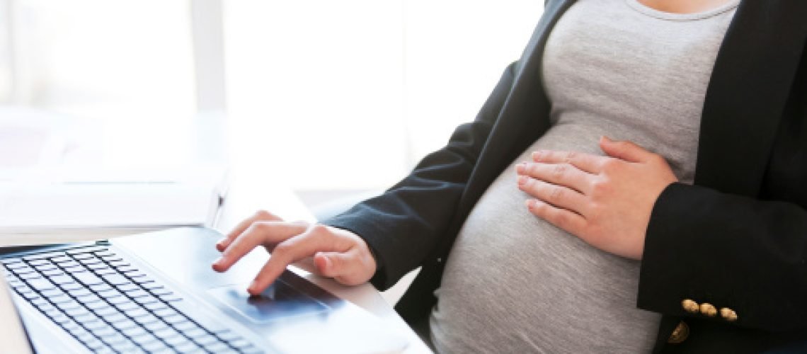 Cropped image of pregnant businesswoman typing something on laptop while sitting at her working place in office